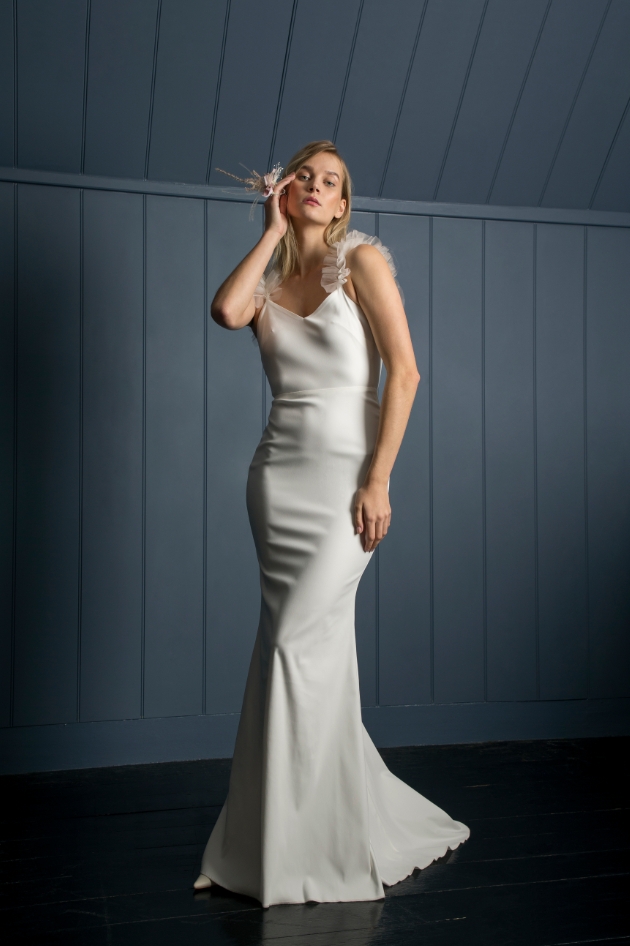 Model in front of wooden blue background wears slimline wedding dress with frill straps