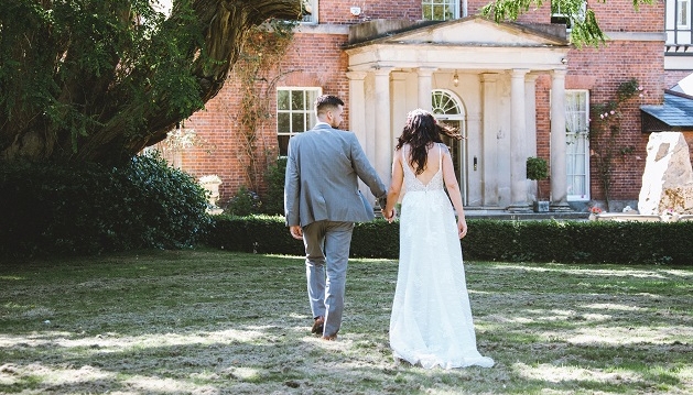 Bride and groom hold hands walking towards a large house