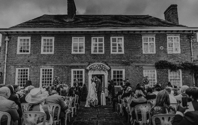 A black and white image of a couple getting married in front of a grand manor house