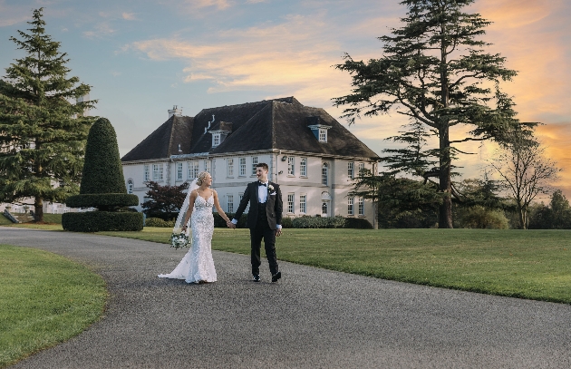A bride and groom walking hand-in-hand outside a white manor house