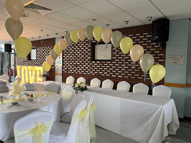 A long white table with a balloon arch above it next to a round table